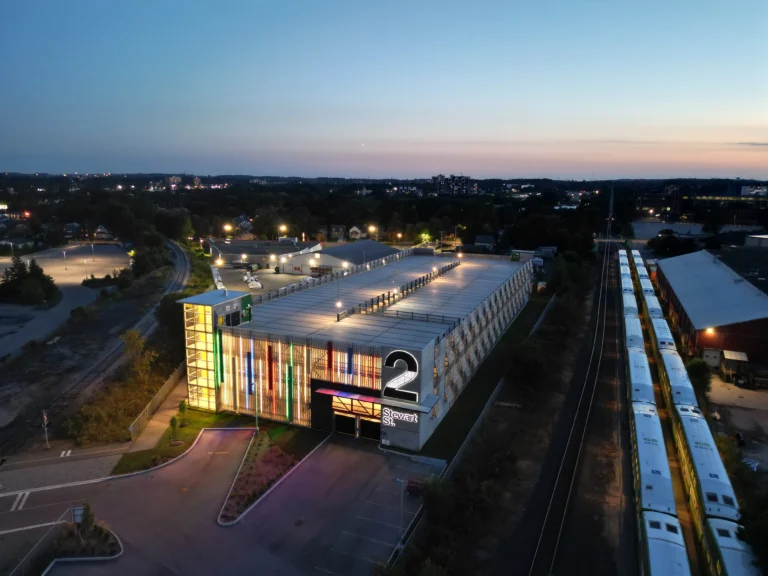 Wider blue-hour aerial of an illuminated multi-storey parking garage with rooftop lights and a brightly colored front facade next to tracks and