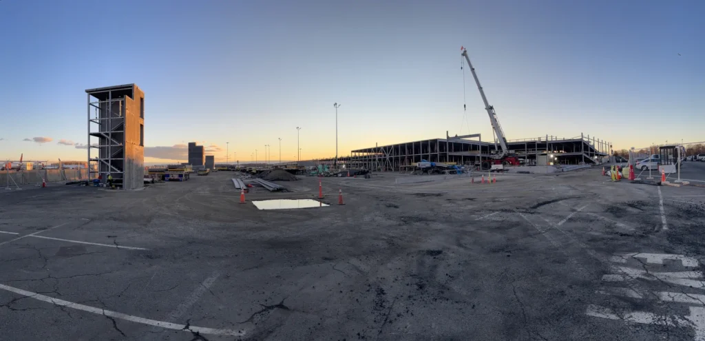 Wide construction-site photograph of a long partly erected parking deck with freestanding stair tower, exposed steel structure, crane and gravel or