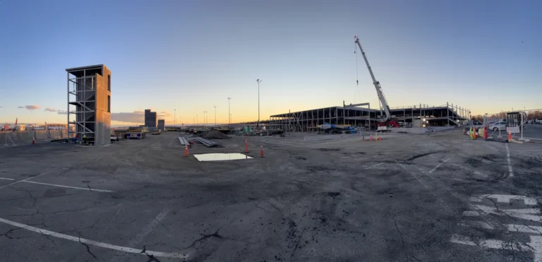 Wide construction-site photograph of a long partly erected parking deck with freestanding stair tower, exposed steel structure, crane and gravel or
